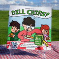A children's book titled 'Dill Chips!' displayed upright on a clear acrylic easel outdoors on a red-and-white checkered picnic blanket. The book cover features cartoon children and a jar of dill chips, with a grassy field and blue sky in the background.