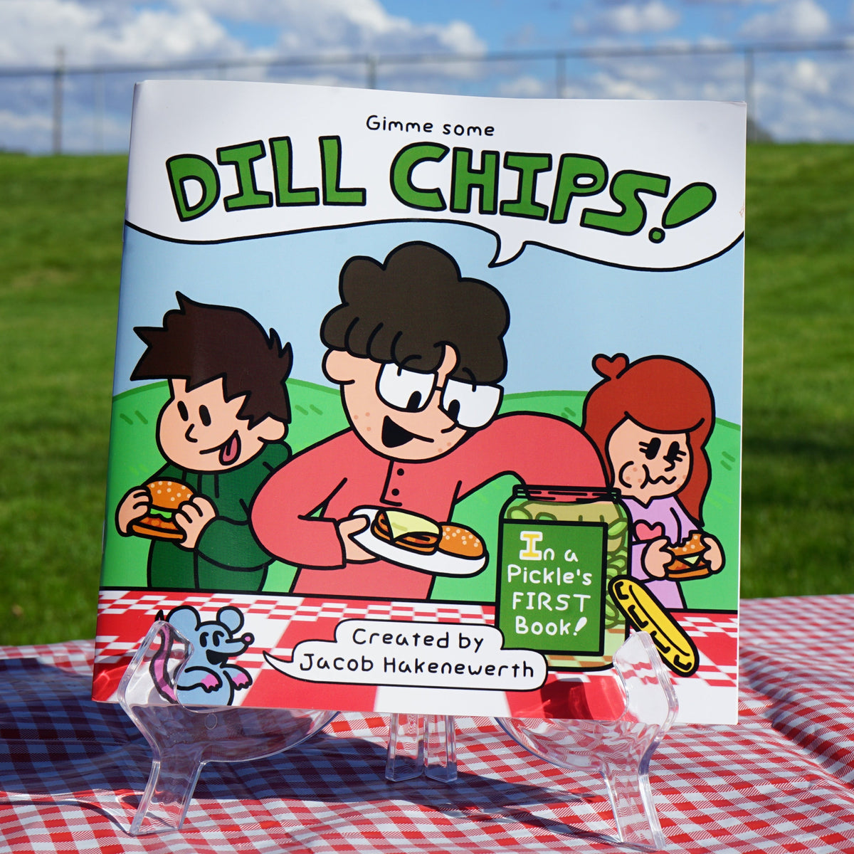 A children's book titled 'Dill Chips!' displayed upright on a clear acrylic easel outdoors on a red-and-white checkered picnic blanket. The book cover features cartoon children and a jar of dill chips, with a grassy field and blue sky in the background.